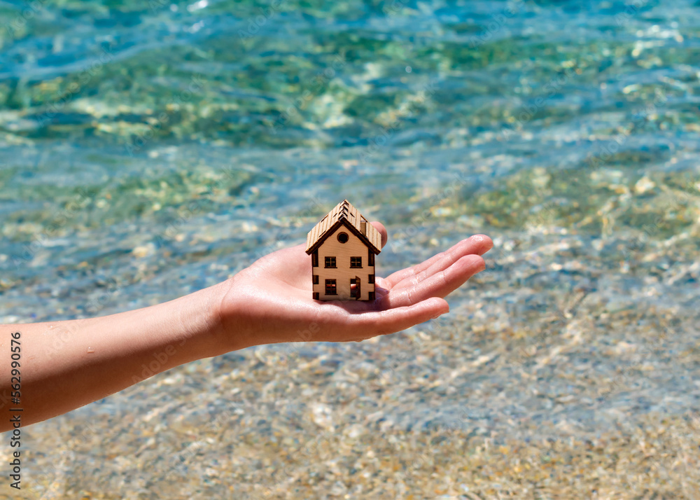 Boy carefull hold model of house above crystal clear water of sea. House in hand of child and summer sea beach. Family holiday property, resort, vacation real estate, save nature as your home