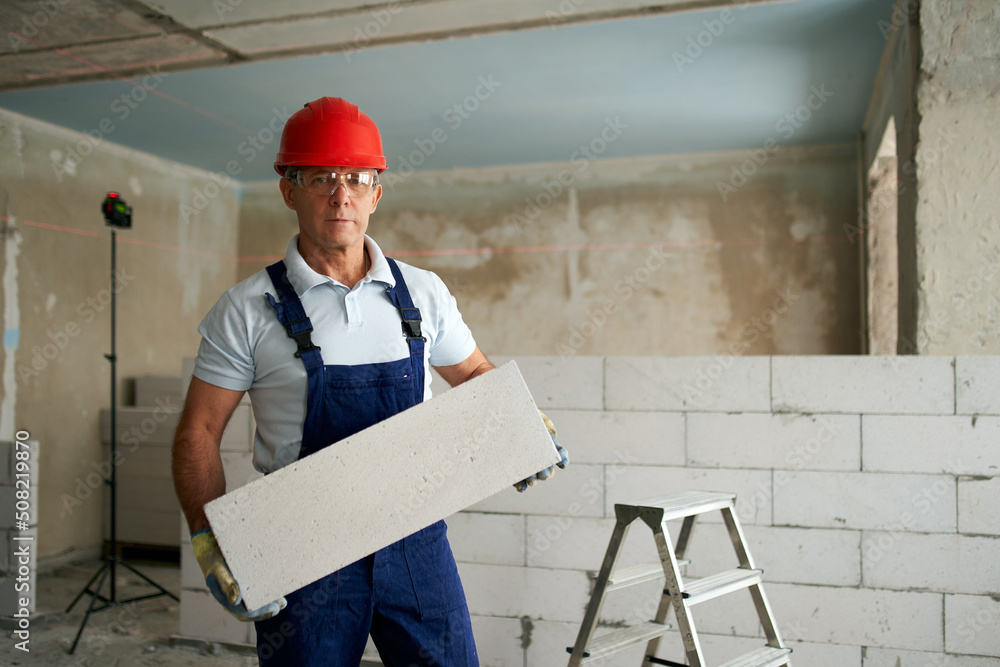 Bricklayer or mason constructs wall of autoclaved aerated concrete blocks. Portrait of brickwork worker contractor standing with quality foamed concrete doing masonry on construction site.